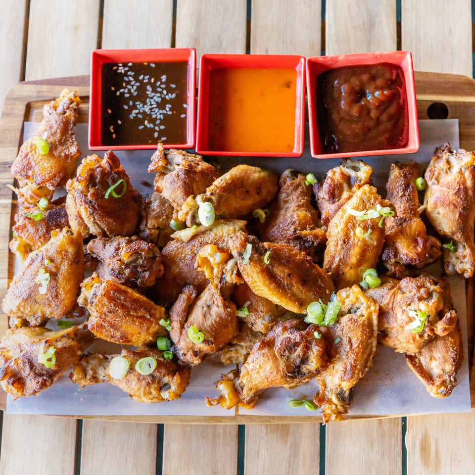 Crispy chicken wings topped with scallions on a wooden serving board, accompanied by three dipping sauces in red bowls.