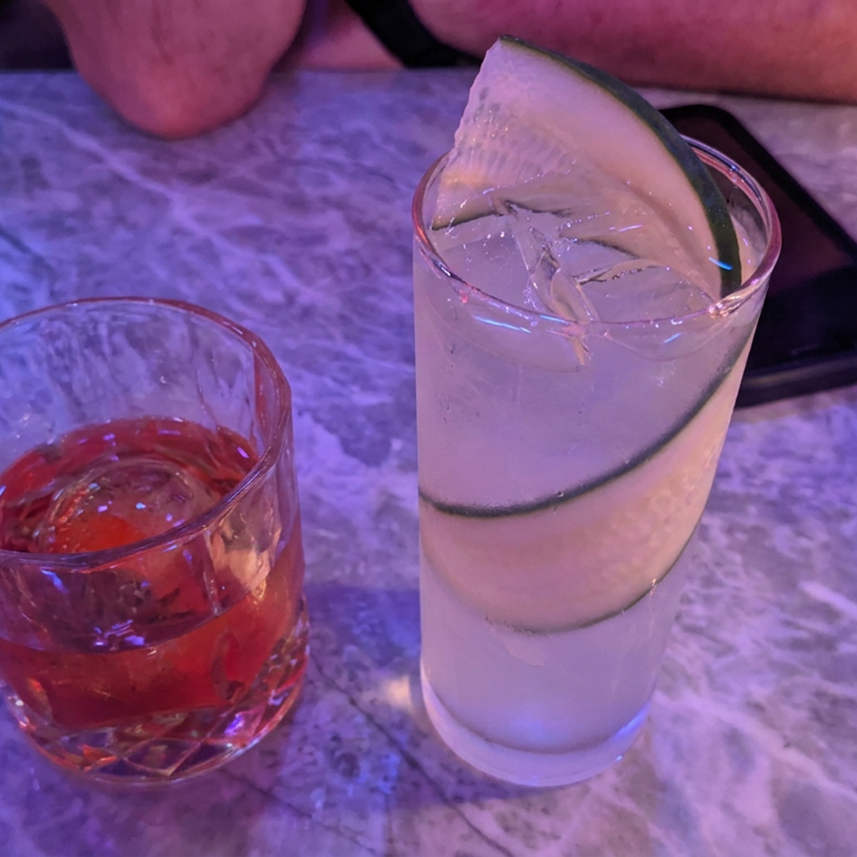 Two cocktails sit on a marble restaurant table: a red drink and a tall citrus drink garnished with lime.
