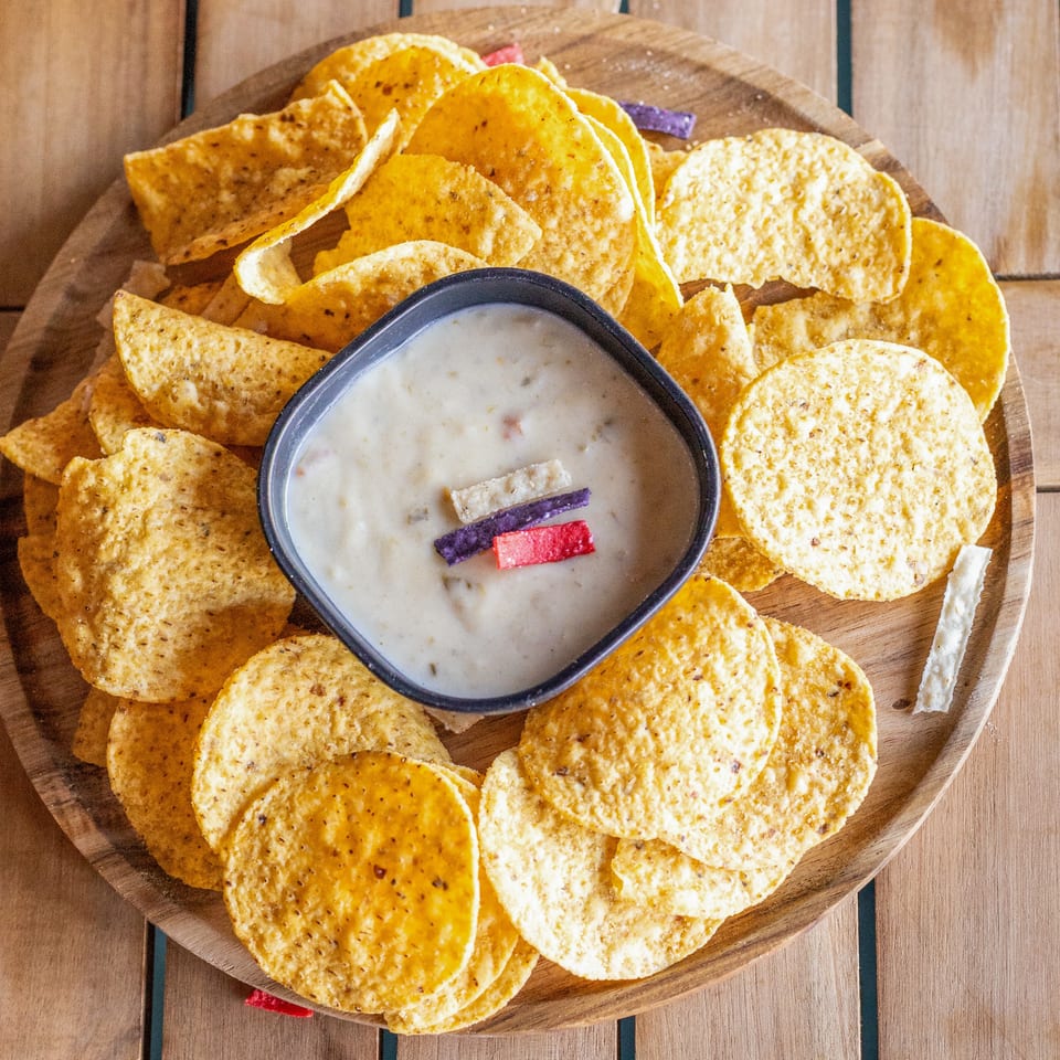 Assorted tortilla chips surround a bowl of creamy queso dip on a wooden serving board.